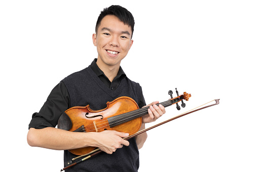 A young male is smiling and looking at the camera, holding up a violin and a bow. He is wearing a black shirt and a vest sweater. He is in a studio with a isolated white background.