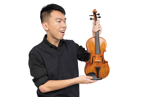 A young male is smiling and looking at a violin that he is holding up. He is wearing a black shirt and a vest sweater. He is in a studio with a isolated white background.