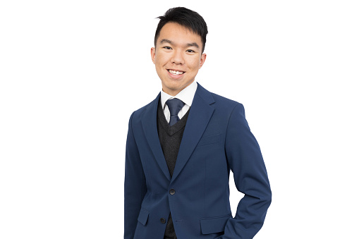 A young male is smiling and looking at the camera, wearing business formal as a businessman. He is wearing a blue blazer, black vest and blue tie. He is in a studio with a isolated white background.