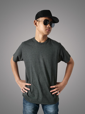 Young Asian teenage boy wearing black shirt, hat and sunglasses standing with hands on hip over grey background, t-shirt template mock up