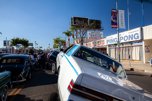 Los Angeles, CA, USA – August 21, 2022: General shot of people at Lowrider event displaying lowriders on three wheels parked outdoors with local business in the background taken from behind cars  in southern California
