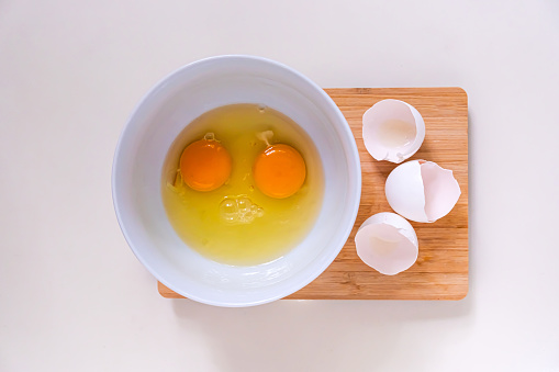 Bright yellow egg yolks in a white bowl and eggshells on a bamboo cutting board.