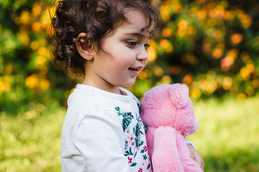 A curly-haired young girl clutches a pink plush toy, lost in thought amidst an autumnal park setting. The warm sunlight enhances the nostalgic and innocent ambiance of this genuine childhood moment.