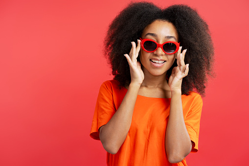 Positive attractive African American young woman with curly hair wearing stylish red sunglasses isolated on red background, copy space, closeup. Advertisement concept