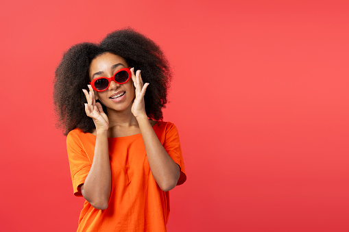 Portrait of smiling beautiful African American young woman with curly hair wearing stylish red sunglasses isolated on red background, copy space. Advertisement concept