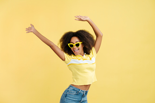 Portrait of beautiful positive African American young woman with curly hair wearing stylish yellow sunglasses isolated on yellow background, closeup. Summer concept