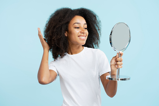 Portrait of beautiful African American woman with curly hair holding mirror looking into it isolated on blue background. Concept of morning routine, hair care