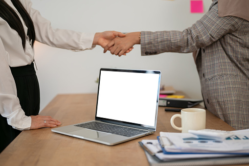 Cropped image of two businesswomen are shaking hands after the meeting in a meeting room. A white-screen laptop computer mockup on a meeting table. dealing business, giving trust, confirmation