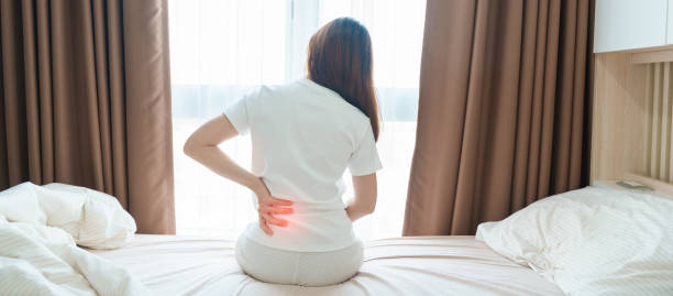 woman having back body ache during sitting on bed at home. adult female with muscle pain after waking up due to piriformis syndrome, low back pain and spinal compression. health medical concept - dor de costas imagens e fotografias de stock