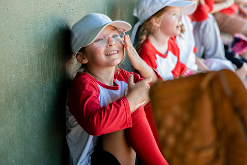 Adorable elementary age girl adjusts her glasses while sitting in dugout with little league baseball team