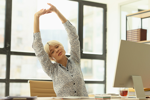 Overworked blonde woman stretches arms and back sitting at table near computer. Tired female secretary warms up body at workplace in office