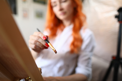 Close-up of red haired woman holding paintbrush with oil paint. Talented female artist wearing white shirt. Process of creating masterpiece. Studio at home. Art concept