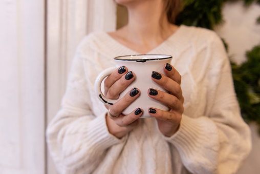 Close-up of female hands with black manicure holding white stylish cup with hot drink. Winter cocoa, coffee from girl sitting by window in white knitted sweater. Warmth comfort atmosphere. Cozy winter
