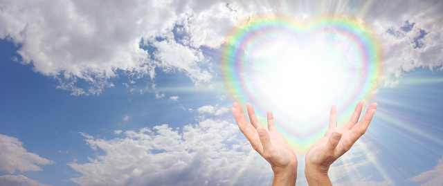 blue sky and fluffy clouds with a heart shaped starlight filled rainbow heart and female hands sending healing energy