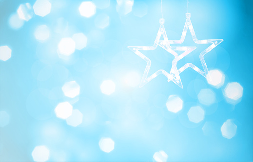 Horizontal illustration of a creative light blue color Xmas lights bokeh shimmery backgrounds with pair of hanging shining pentagram stars. It is textured and has a color gradient. There is no text and no people, ample copy space. Apt for celebrations backdrops, wallpapers, templates for greeting cards, banners or posters or gift wrapping paper sheets.