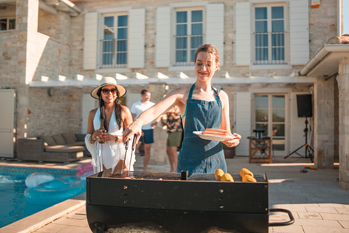 At a villa pool party, a delightful Caucasian woman enjoys herself while grilling food for a diverse group of friends, creating a memorable summer scene.