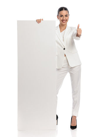 sexy elegant woman in white suit holding empty board and making thumbs up gesture while smiling on white background