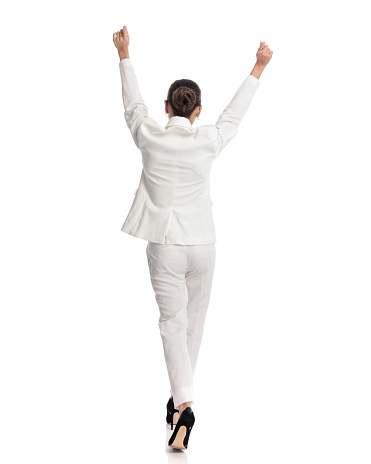 back view of elegant woman in white suit holding arms above head and cheering while walking in front of white background