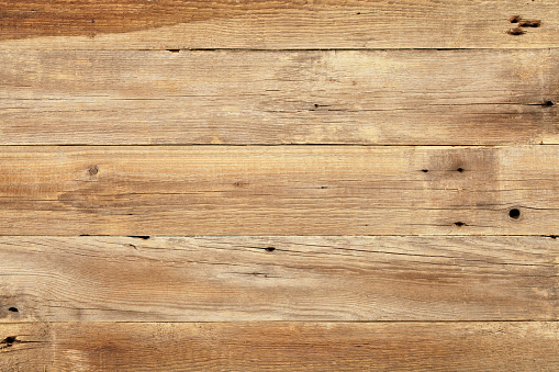 A close-up view of the texture of a wooden table.  The table is made from light-colored wood and has some dark spots on the left and right sides.  There is also a dark spot in the middle of the table toward the bottom.  The wood at the top and bottom of the table is darker than the wood in the center of the table.  The piece of wood at the top of the table also appears to have a dark stain on it.  The close-up gives a direct view from above the table, revealing cracks and knotholes in the wood.