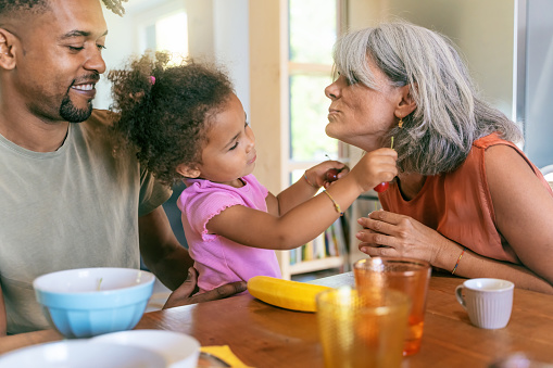 A biracial child joyfully interacts with her grandmother using cherries as earrings, while her African father observes with mixed emotions.