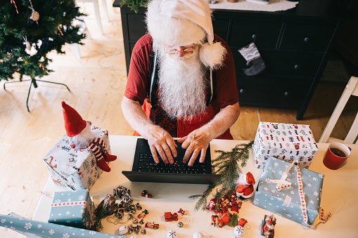 Santa Claus takes a moment to connect with the digital age, working on a laptop in a room filled with the warm glow of Christmas lights and a beautifully decorated tree.