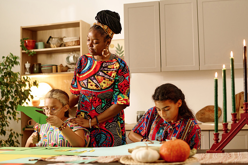 Young woman in national attire standing by her daughter cutting green paper while making greeting card for kwanzaa celebration at home