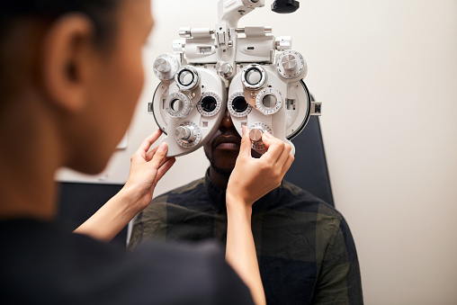 Optometrist, patient and phoropter in clinic, help or lens for check, eye exam or healthcare for wellness. Ophthalmologist, person and machine to test eyesight, care or vision for results in hospital