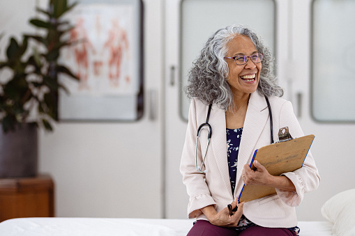 Portrait of a vibrant senior adult female doctor of Hawaiian and Chinese descent with grey hair smiling as she sits in a medical examination room and waits for a patient.