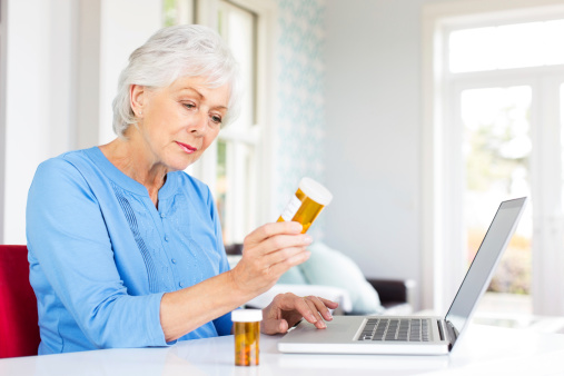 Senior woman with pill bottles researching on laptop at home. Horizontal shot.