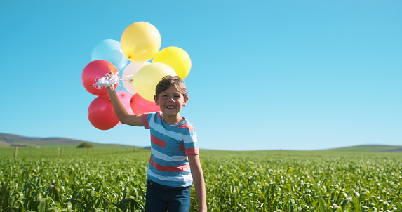 Child, field and holding balloons in countryside by sustainable farm with happiness, energy and excited in sun for adventure. Nature, plants and fashion boy with fun freedom, agriculture and blue sky