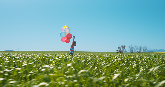 Child, freedom and balloons in landscape with blue sky in countryside, field or running in environment. Happy birthday, celebration and kid with balloon in the air with meadow, grass and nature