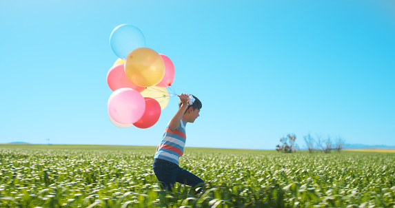 Child, field and run with balloons in countryside, birthday adventure and happiness with energy for excited celebration. Nature, plants or young boy with fun in freedom, wellness or happy by blue sky