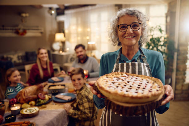 Happy senior woman with Thanksgiving pie looking at camera. Her family is in the background.