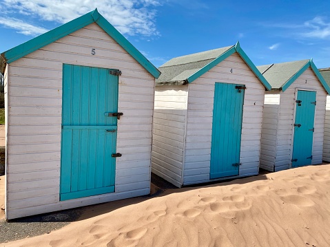 Row of three beach huts, all with blue doors. A mound of sand has blown up against them. Bright blue sky on a sunny day
