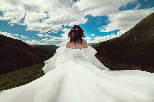 The bride turns her back and runs away in a white dress with flowing hair in a green valley against the backdrop of mountains and hills. A special day for a woman.