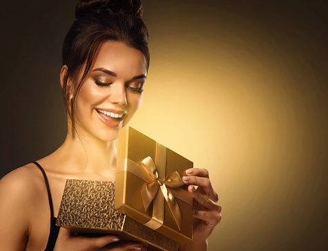 Gift giving for Woman. Beautiful Girl opening Present Box with shining Light. Happy smiling Model receive Surprise over Golden background