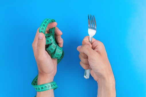 Fork and tape measure in male hands on a bright blue background. A man with a hand tied with a measuring tape holds a fork close-up.