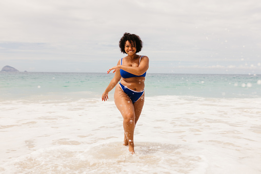 Happy young woman celebrating the spirit of summer on the beach, dancing joyfully in her bikini. Body confident, plus size woman having fun on a solo beach holiday.