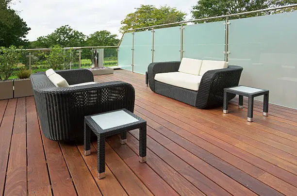 the wooden decking at the rear of a luxury apartment with garden views in the background. Two large outdoor settees with weatherproof cushions are set opposite each other with coffee tables on the side. A large glass and stainless steel division can be seen to the right with clear glass at the rear.
