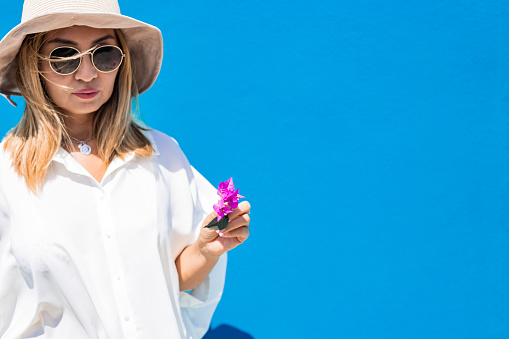 attractive young blonde asian woman in sunglasses and a hat in white oversize shirt dress against blue wall background isolated with copy space