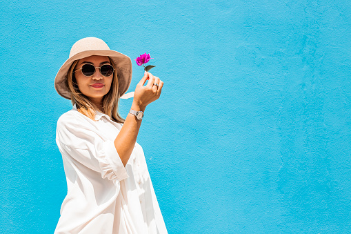 attractive young blonde asian woman in sunglasses and a hat in white oversize shirt dress against blue wall background isolated with copy space