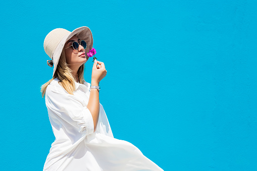 Portrait of a beautiful young asian woman in sunglasses, white dress and hat on blue background smells a pink flower. copy space