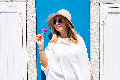 Portrait of a beautiful young asian woman in sunglasses, white dress and hat on blue background smells a pink flower. copy space