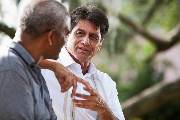 Multi-ethnic men talking Multi-ethnic men (50s and 60s) sitting outdoors, having conversation.  Focus on man in white shirt (Hispanic/Native American, 50s). serious talk stock pictures, royalty-free photos & images