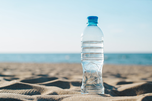 Plastic bottle of water standing on beach at sea. Refreshing cold drink on hot sunny day, copy space.