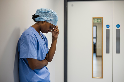 Side view waist up of a female doctor standing in the hospital corridor. She's dressed in medical scrubs and she has her back against a wall, she has her head down and her hand on her head, she's feeling concerned in a hospital in Newcastle, England.
