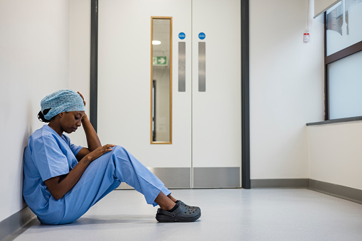 Full length side view of a female doctor sitting on the floor in the hospital corridor. She's dressed in medical scrubs and she has her back against a wall, she has her head down and her hand on her head, she's feeling concerned in a hospital in Newcastle, England.