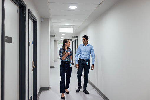 Front view full length of two doctors having a conversation in a hospital corridor, wearing button-down shirts. They discuss patient care, reflecting their dedication and expertise. The scene exudes professionalism and trust in their collaborative approach to medicine in a hospital in Newcastle, England.