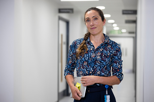Waist up of a female doctor standing in the hospital corridor. She's dressed in a button-down shirt and has a warm smile on her face and she's holding a digital tablet, she looks directly at the camera, radiating professionalism and a welcoming demeanour.