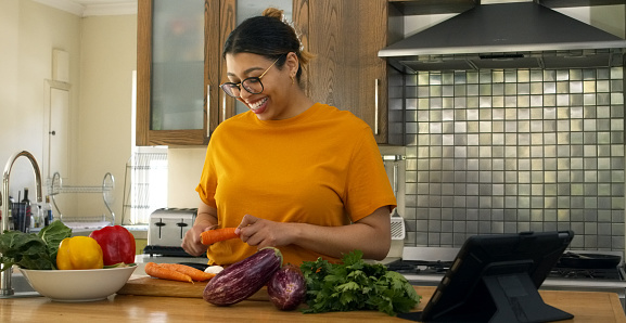 Woman, smile and cutting carrot by tablet, tutorial or vegan nutrition in home kitchen for meal prep. Chef girl, food and live stream for diet, healthy fruits or leaves with knife for cooking process